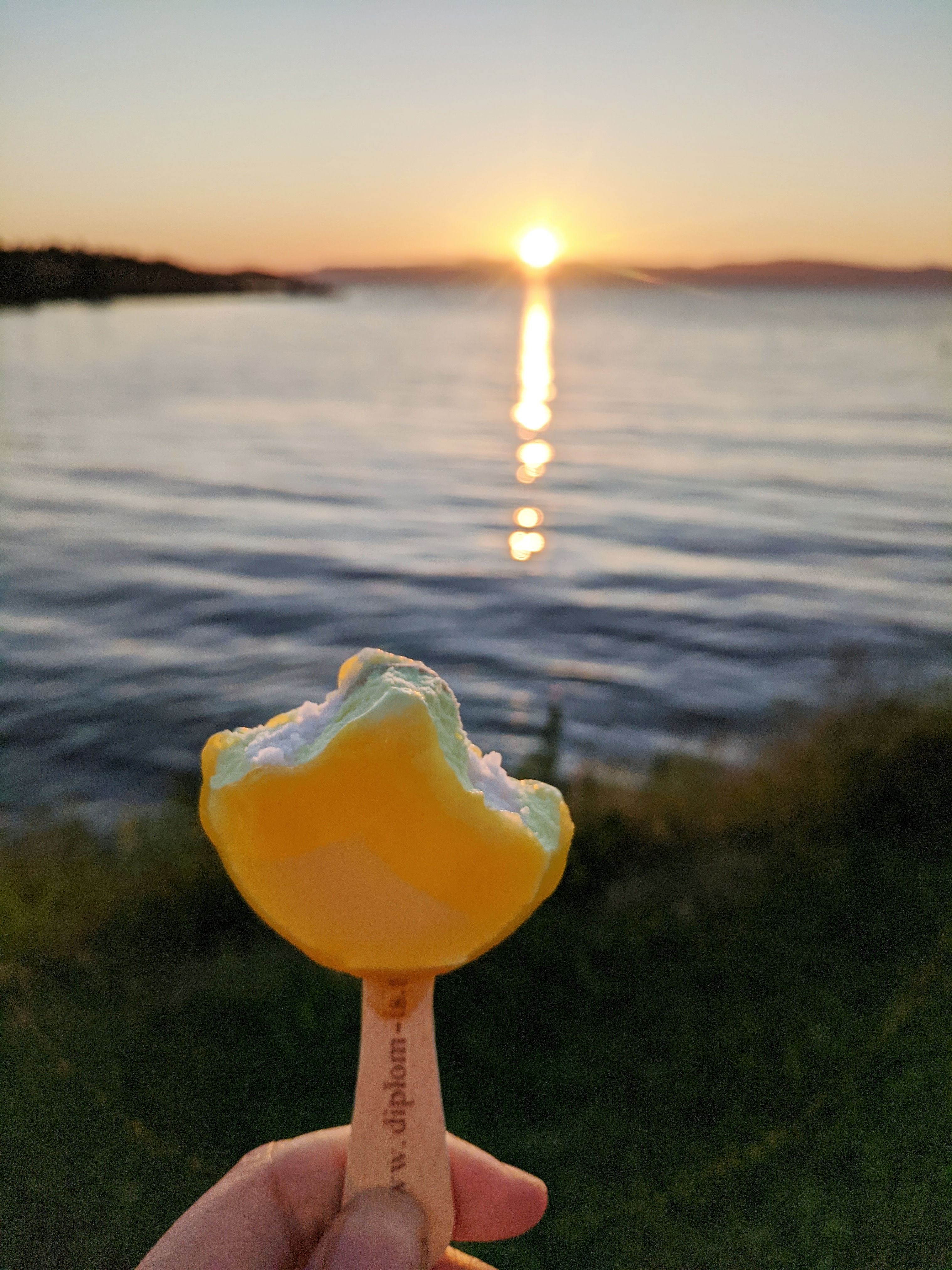 A half-eaten lemon popsicle is held up in front of the Trondheim fjord at sunset