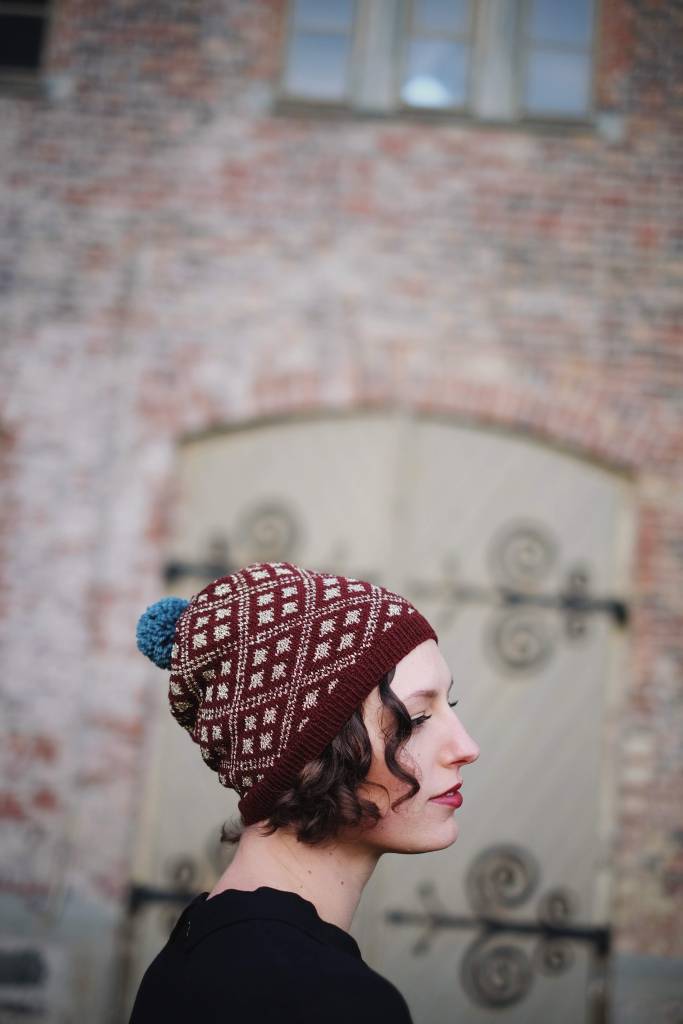 Dianna wears a burgundy and gold hat with a pattern of colorwork diamonds while standing in front of an old brick wall and set of doors with fancy ironwork.