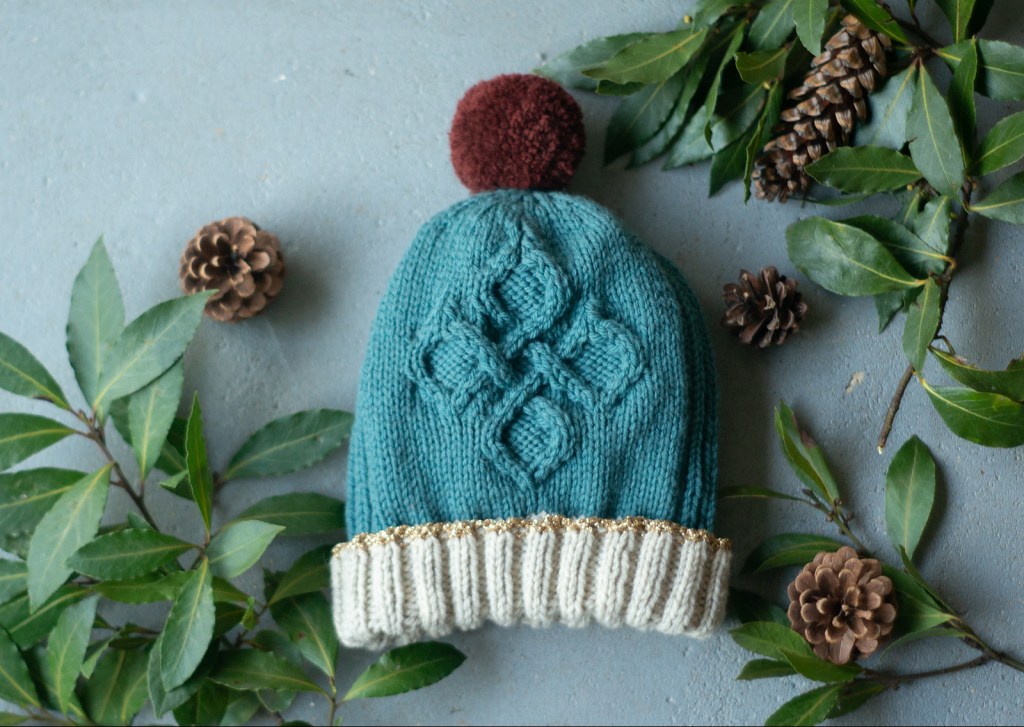 A knitted hat featuring a diamond cable pattern in tealy blue with off-white trim and a burgundy pom pom lays on a grey floor surrounded by leafy branches and pine cones.