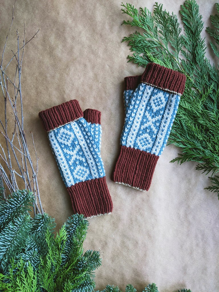 A pair of colorwork fingerless mitts in shades of burgundy, off-white, and blue laid out on brown paper with evergreen branches arranged around them.