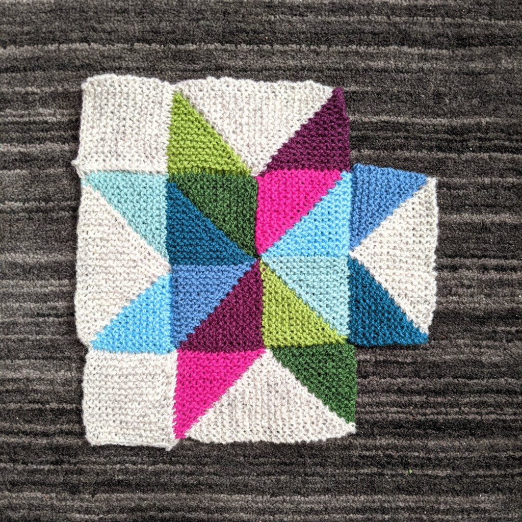 A multi-colored eight-pointed star knitted in garter stitch lays on a dark grey carpet.