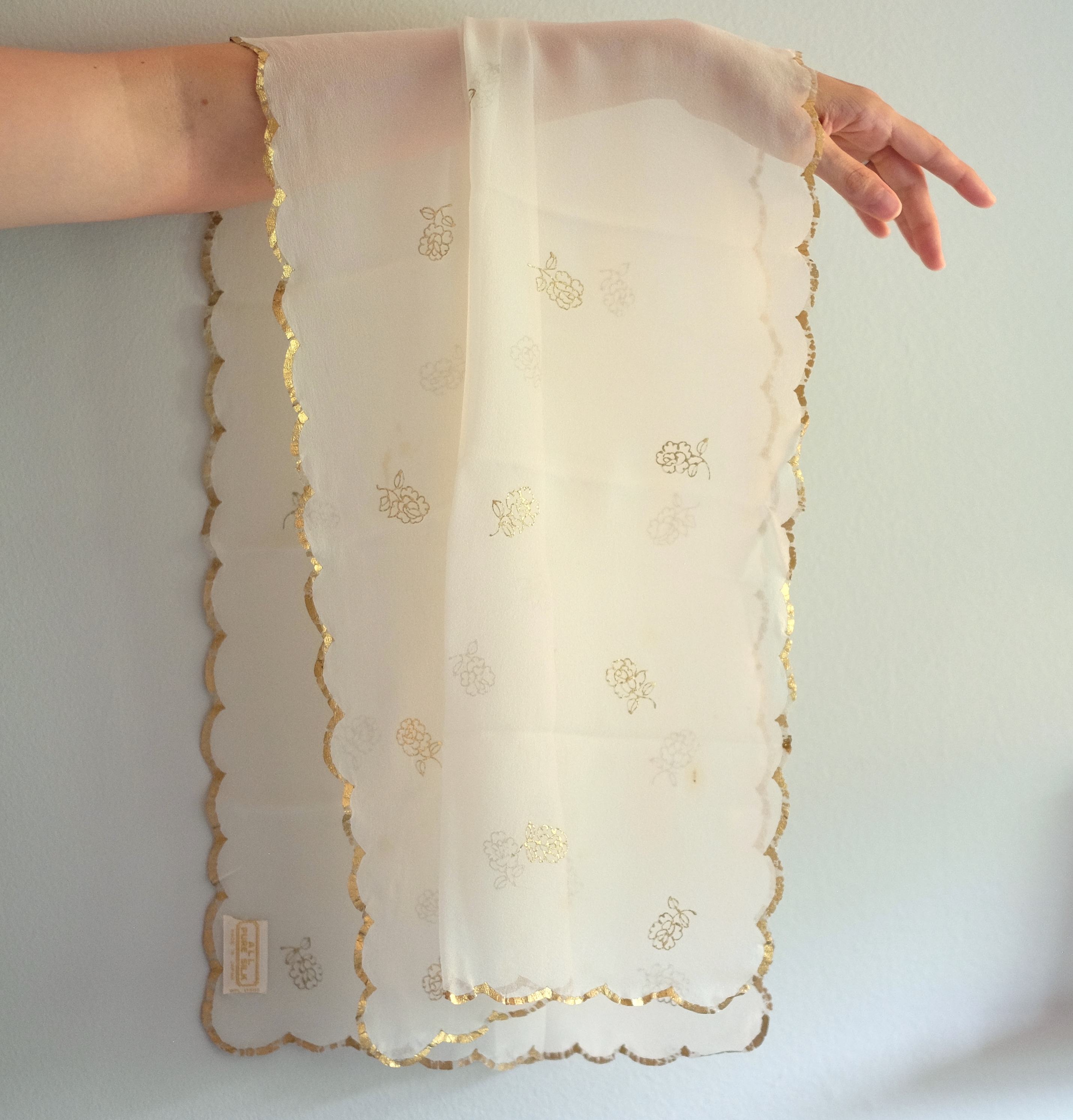 A silk scarf hangs over an extended arm. The scarf is off-white and translucent, with a pattern of flowers printed in gold and a gold scalloped edge all the way around.