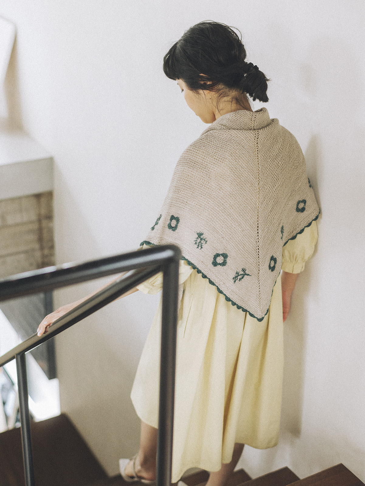 A dark-haired woman faces away from the camera as she walks down a set of stairs. She wears a pale yellow dress and a triangular shawl is draped over her back and shoulders. The shawl is a beige color with an embroidered border of flowers and branches in dark green, and a scalloped