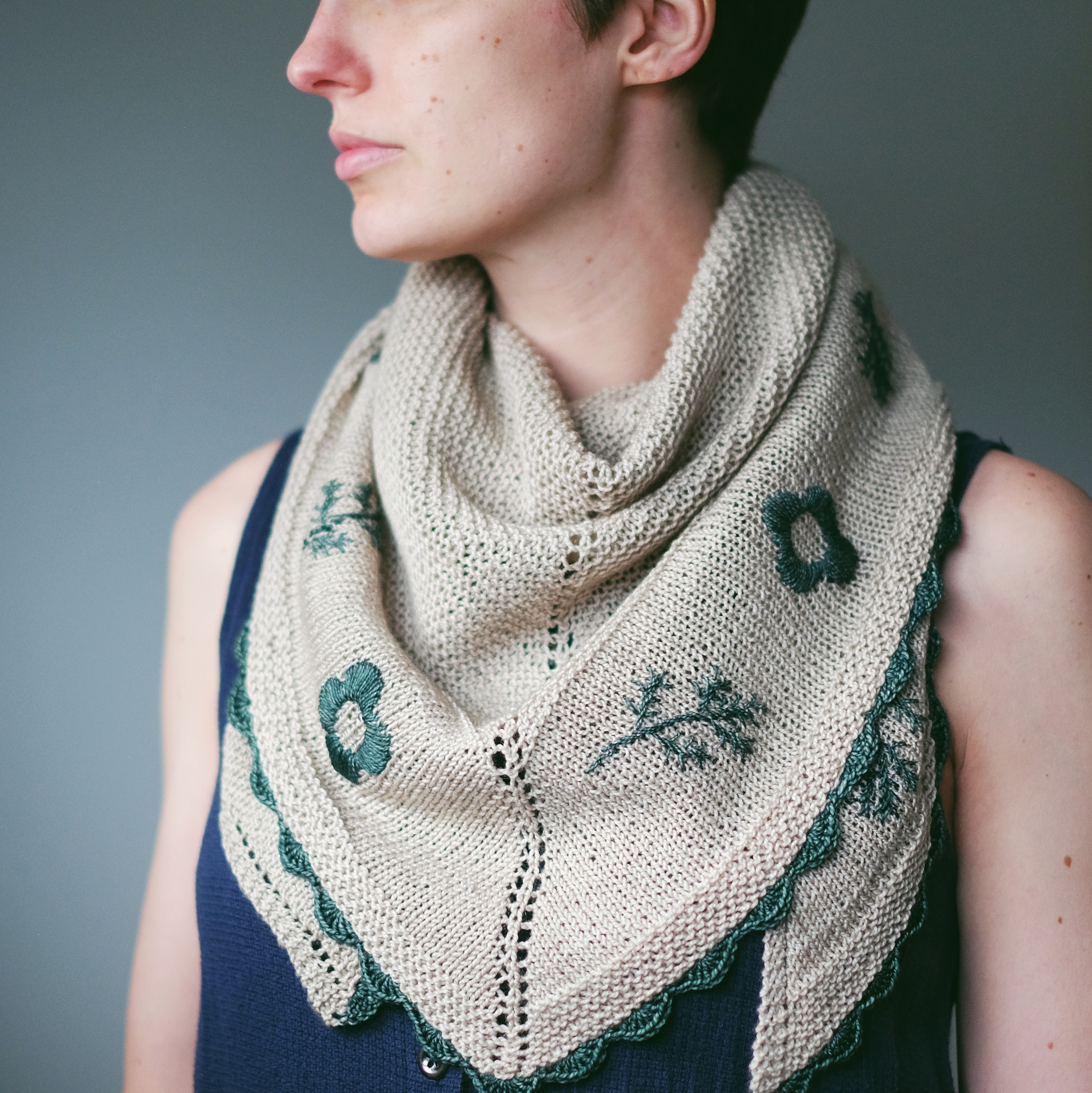 A woman with a short pixie cut wears the triangular shawl kerchief-style around her neck over a sleeveless dress. The dark green embroidery shines in the light.