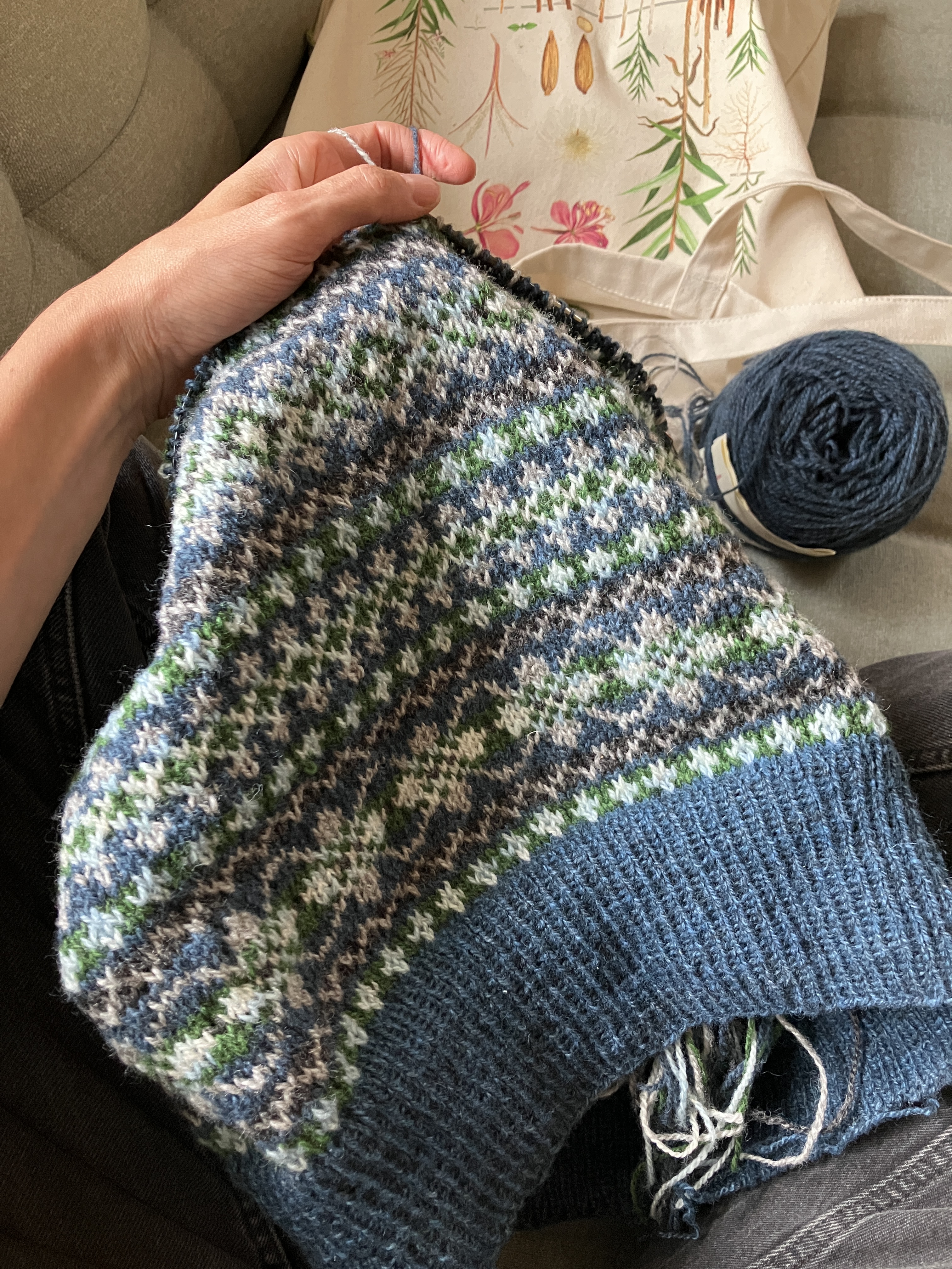 A knitted fair isle style vest in progress, sitting in Dianna's lap. The motifs are in shades of indigo blue, green, and grey. A ball of dark blue yarn sits to the right and two strands of yarn (light and dark blue) are held over Dianna