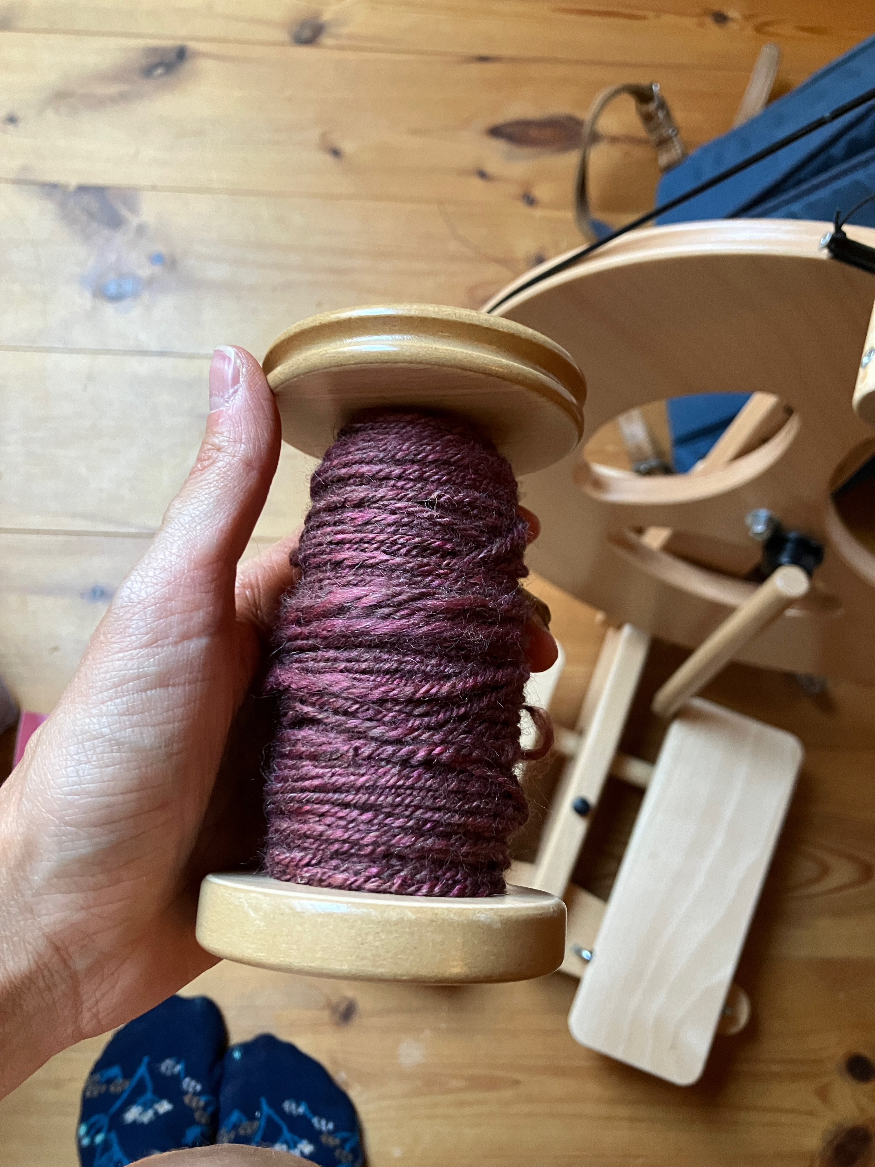 Dianna holds a wooden bobbin full of freshly chain-plied handspun in pinky-burgundy wool. The Louet spinning wheel used to ply sits slightly out-of-focus below the bobbin.