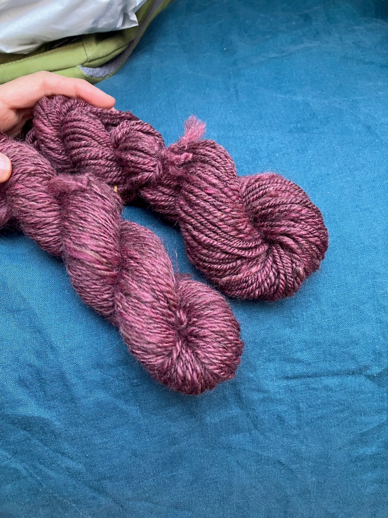 Two skeins of pinky-burgundy chain-plied handspun lay on a petrol blue bedspread. The amount of twist is noticeably different, with the one on the right looking slightly over twisted and the one on the left looking slightly under twisted.