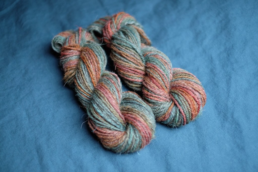 Two skeins of handspun yarn lay side by side on a petrol blue bedspread. They are multi-colored and chain-plied to maintain the color shifts throughout the skeins (so there's no marling). The palette is a mix of teals, pinks, oranges, and some green peeking through.