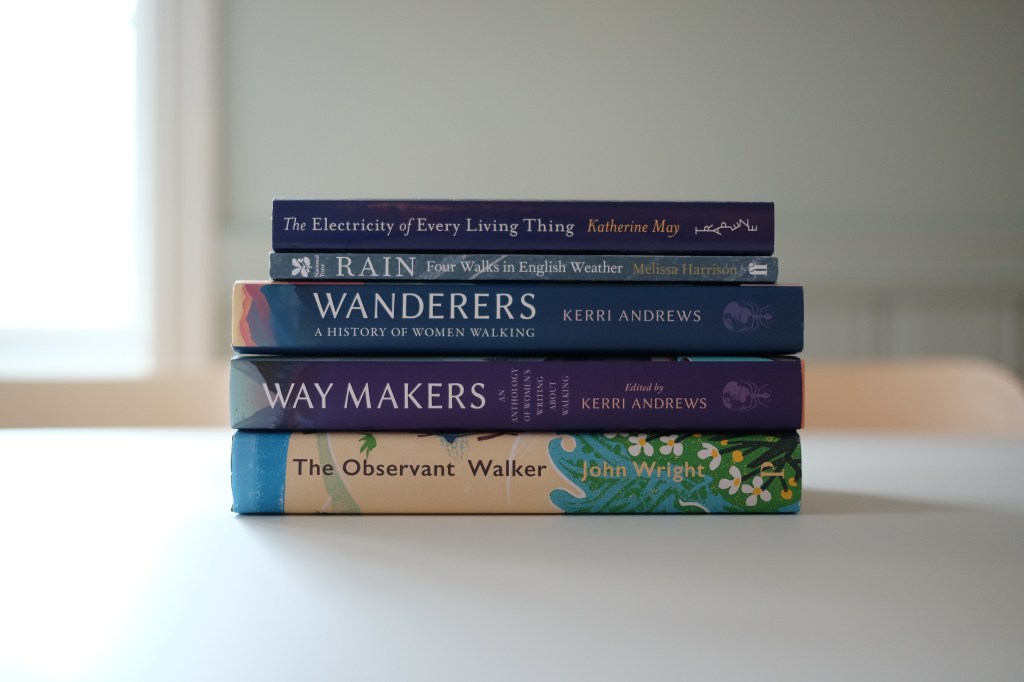 A stack of books sits on a white tabletop, spines visible. From top to bottom, the titles and authors read: The Electricity of Every Living Thing by Katherine May, Rain: Four Walks in English Weather by Melissa Harrison, Wanderers: A History of Women Walking by Kerri Andrews, Way Makers: An Anthology of Women's Writing About Walking edited by Kerri Andrews, and The Observant Walker by John Wright.