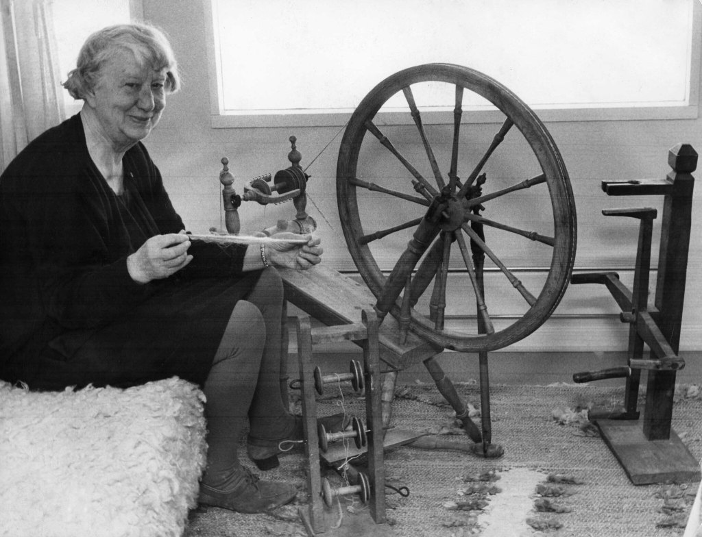 An older woman sits next to a large saxony-style spinning wheel. She is stretching a length of fiber in her hands but not actually spinning it.