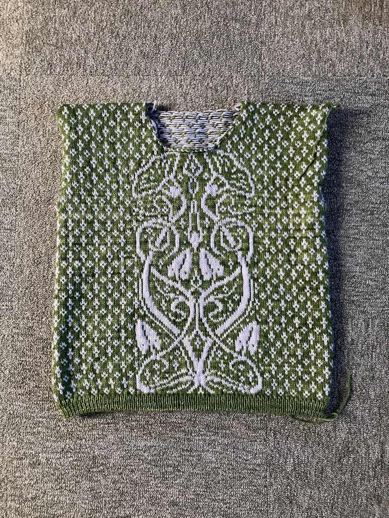 The body of an incomplete colorwork sweater lays flat on a grey-green carpet. The sweater is bright grassy green with allover colorwork in a pale blue, and a central panel featuring bindweed in an art nouveau style.