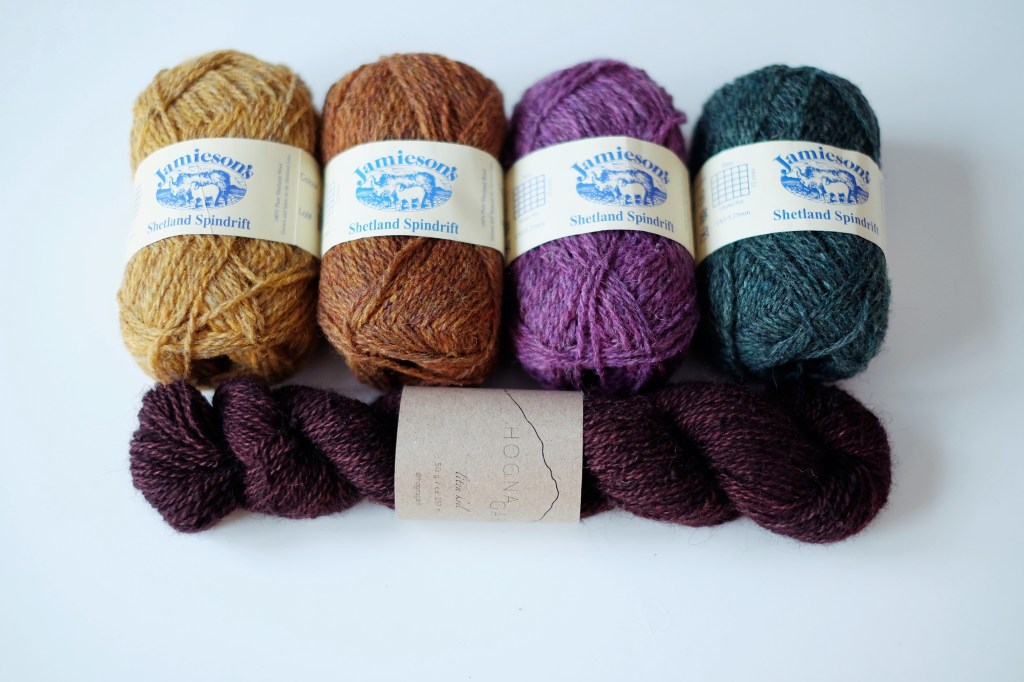 Four balls of Jamieson's Shetland Spindrift yarn lay on a white tabletop above a twisted hank of Hogna Garn. The Jamieson yarns are an ochre yellow, a rusty orange, an orchid purple, and a cool forest green. The Hogna Garn is a purply brown.