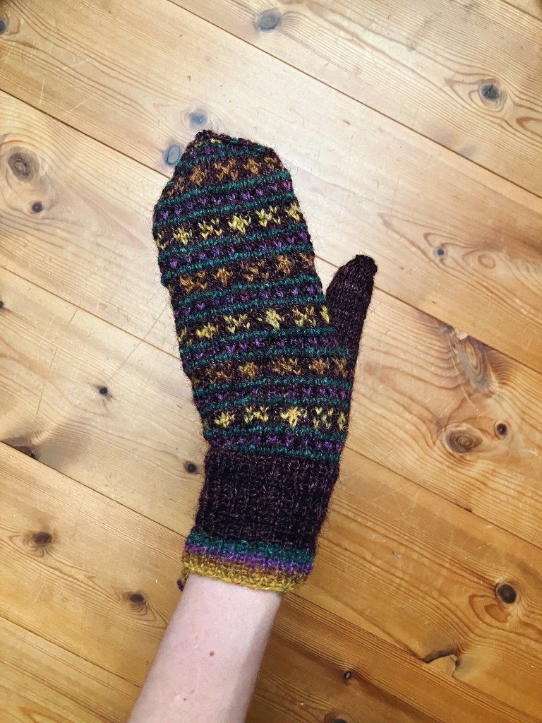 A mitten being modeled in front of a wooden floor. The main color is a deep purply brown, and peerie X and O motifs alternate in shades of ochre yellow, rusty orange, orchid purple, and a cool forest green.