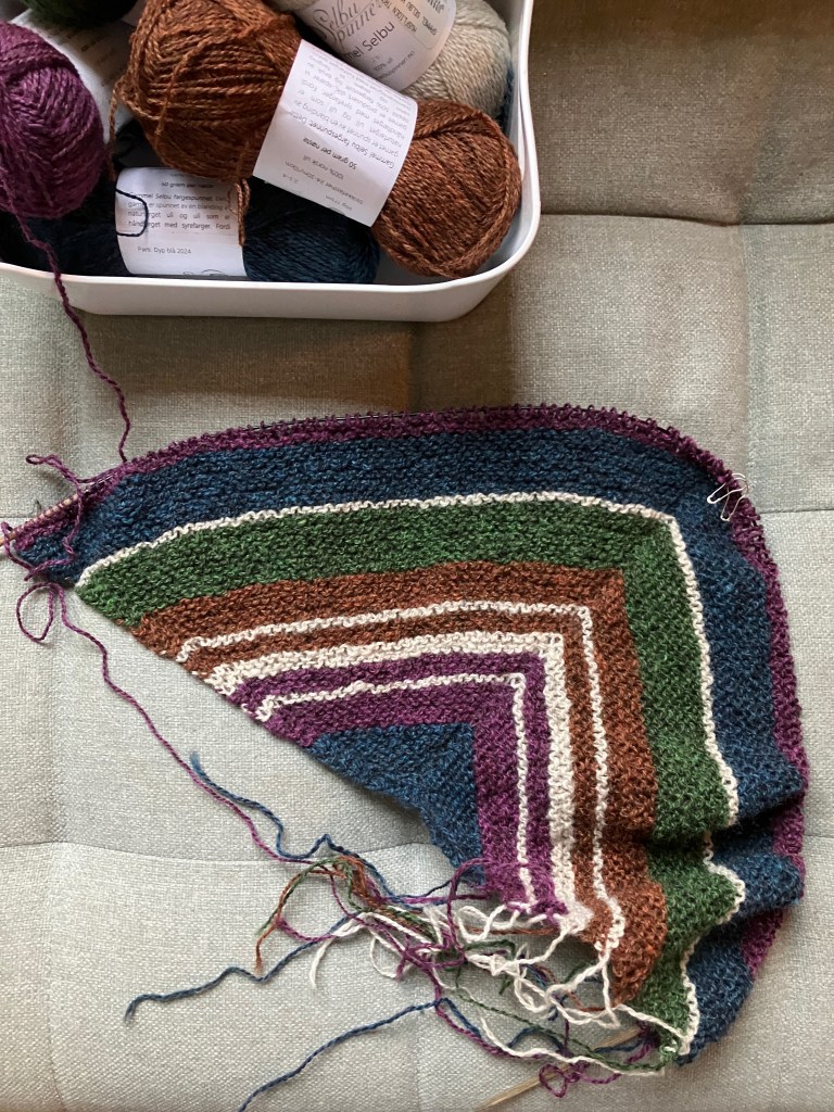 A shawl project in progress, seen from above. A garter stitch triangle featuring stripes of different widths in blue, purple, orange, green, and light grey lays next to a small bin containing the skeins of yarn in the same colors.