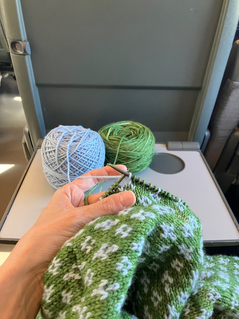 A knitting project on a train. A green and light blue color work sweater is being worked on in a train seat. The two balls of yarn sit side by side on the fold-down tray table.