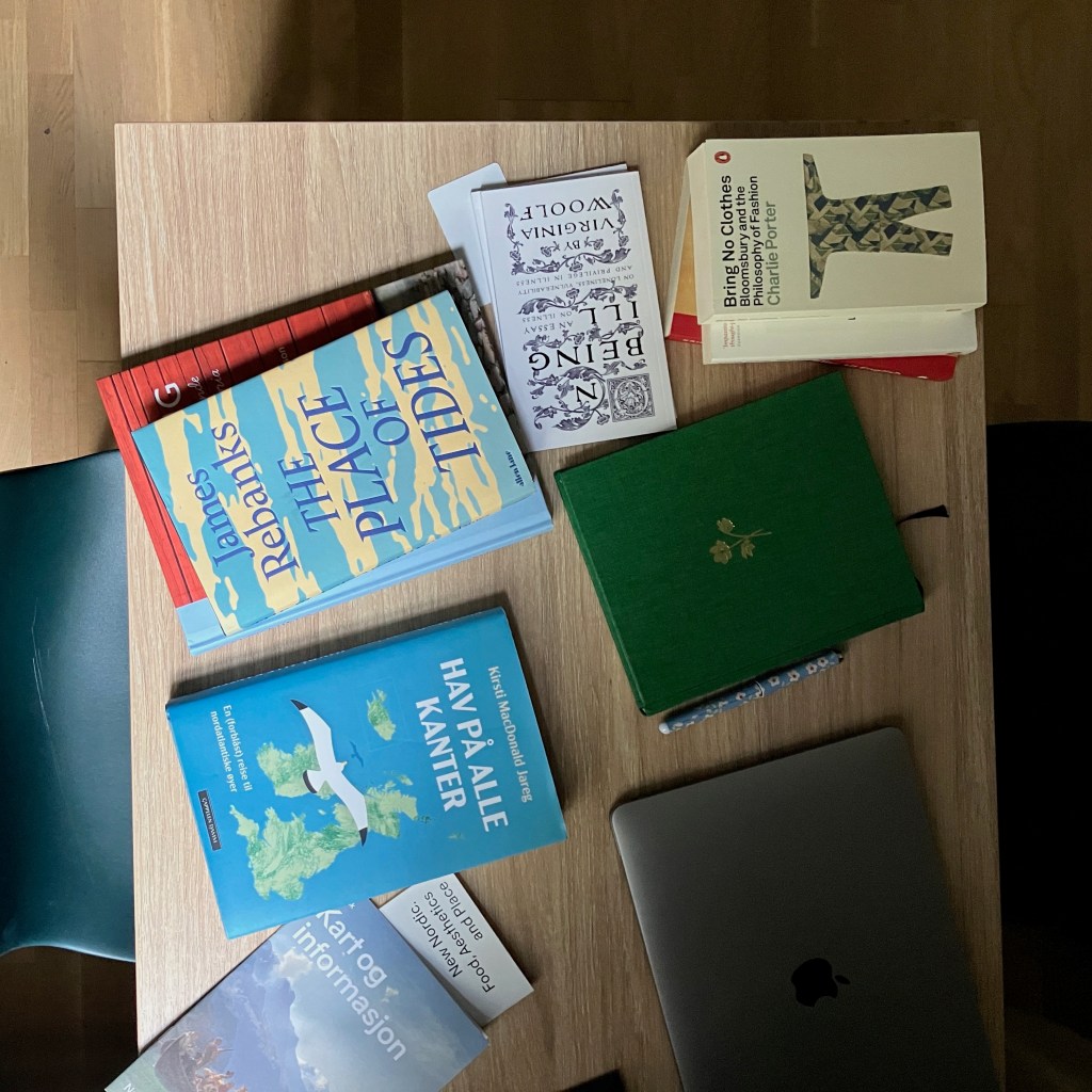 Several piles of books laying on a kitchen table, seen from above. Visible titles include "Hav på alle kanter" by Kirsti Macdonald Jareg, "Place of the Tides" by James Rebanks, "On Being Ill" by Virginia Woolf, and "Bring No Clothes," by Charlie Porter.
