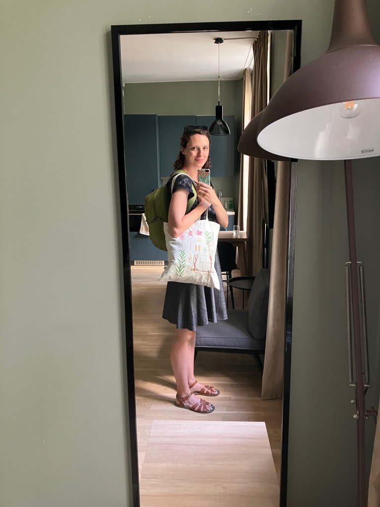 A mirror selfie of Dianna in her hotel room. She turned to one side and smiling at the camera. She wears a backpack and holds a tote bag over one shoulder.