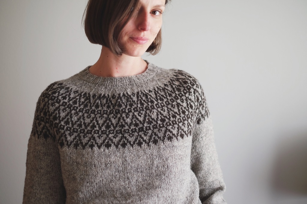 Dianna, a white woman with a brown chin-length bob, smiles whole modeling a newly finished sweater in light grey with a dark brown color work yoke. The yoke is three rows of a zig-zagging pattern based on Latvian embroidery.