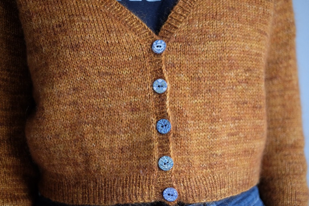 The buttoned front of a cropped V-neck cardigan, being worn. Five buttons feature Morris & Co. designs in blue tones.
