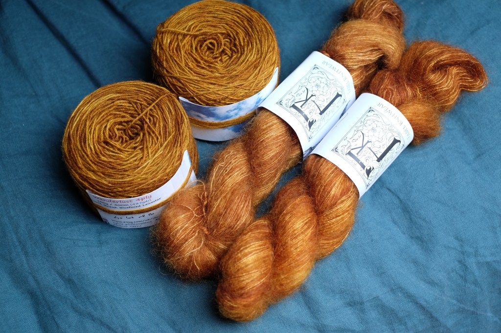 Two caked balls of yellowy brown yarn and two twisted hanks of mohair silk yarn in a similar color lay on a petrol blue duvet cover.