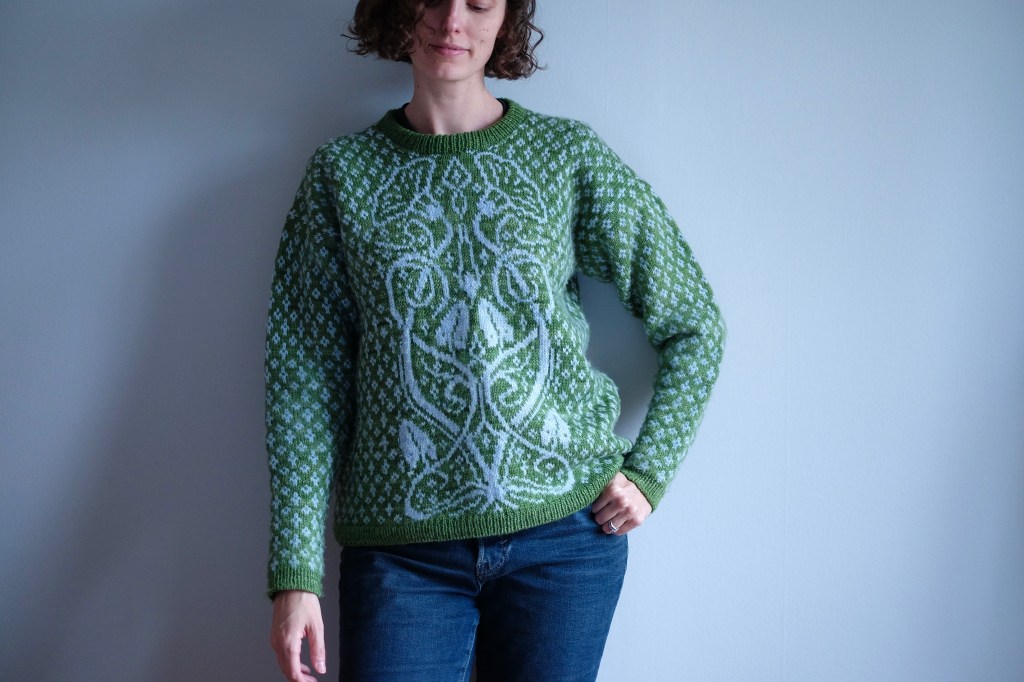 Dianna wears an allover colorwork pullover with a pair of jeans, leaning against the wall with one hand in her pocket. The sweater is grassy green with light blue colorwork, featuring a central panel of bindweed in an art nouveau style.