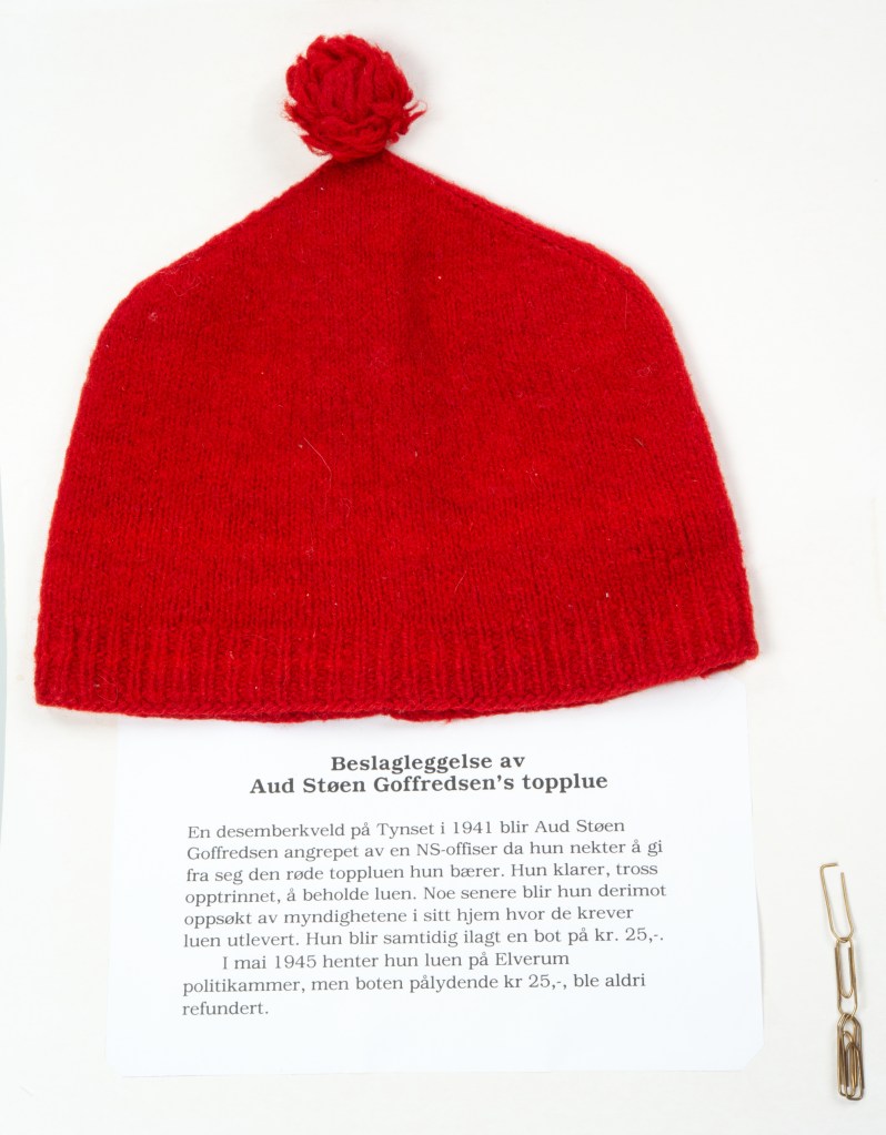 A bright red knitted hat, slightly pointed with a red pom pom on top, lays flat in a museum display case. Several gold paper clips lay in the corner. A description of the hat's history is printed on paper (see description which follows the image).