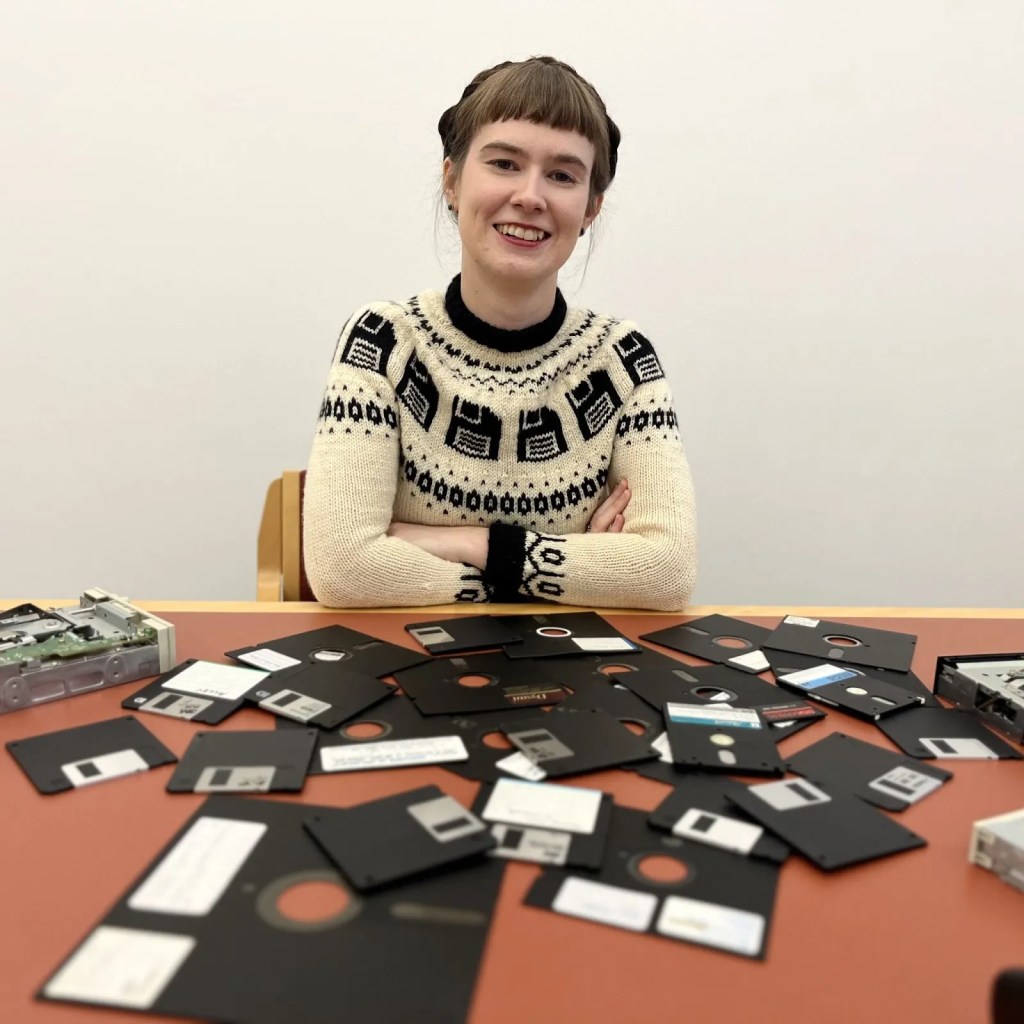 Leontien Talbloom sits at a table strewn with floppy disks. She wears an off-white sweater with a round yoke patterned with black floppy disks.