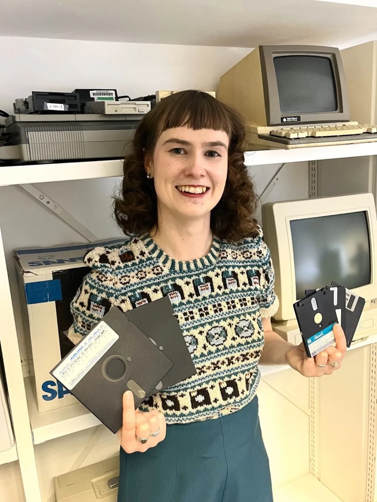 Talbloom holds floppy disks of various sizes while wearing a hand-knitted jumper featuring floppy disks, CDs, and USB sticks. She is standing in front of a shelf with old computers and floppy disk drives.