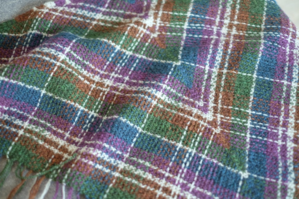 Detail of the tartan pattern with the shawl laying flat on a couch. The colors blend together almost seamlessly, and the yarns that have been vertically woven through the garter stitch are clear to see.