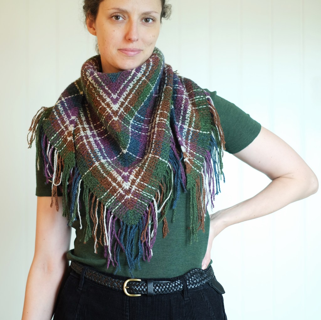 Dianna wears a knitted tartan shawl kerchief-style around her neck and shoulders. The shawl is deep jewel toned colors: green, blue, purple, and orangey-brown, with stripes in a light grey-white.