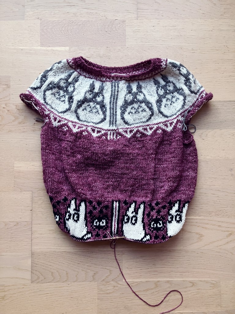 A knitted sweater in progress. It's a purple sweater with a round yoke. The yoke pattern has a white background and a series of grey Totoros around it. At the bottom of the body are white Totoros alternating with soot sprites. The sweater is still on the needles and there are no sleeves yet.