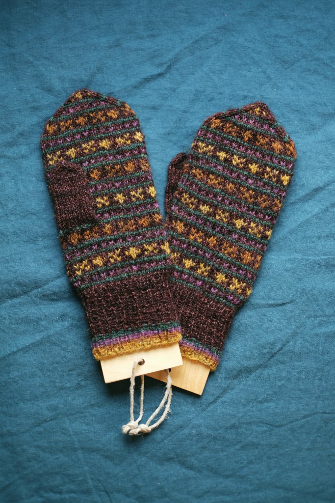 A pair of hand-knitted mittens on wooden mitten blockers laying on a blue bedspread. The main color is a dark purple-brown, with peerie XO motifs in yellow, orange, purple, and forest green.