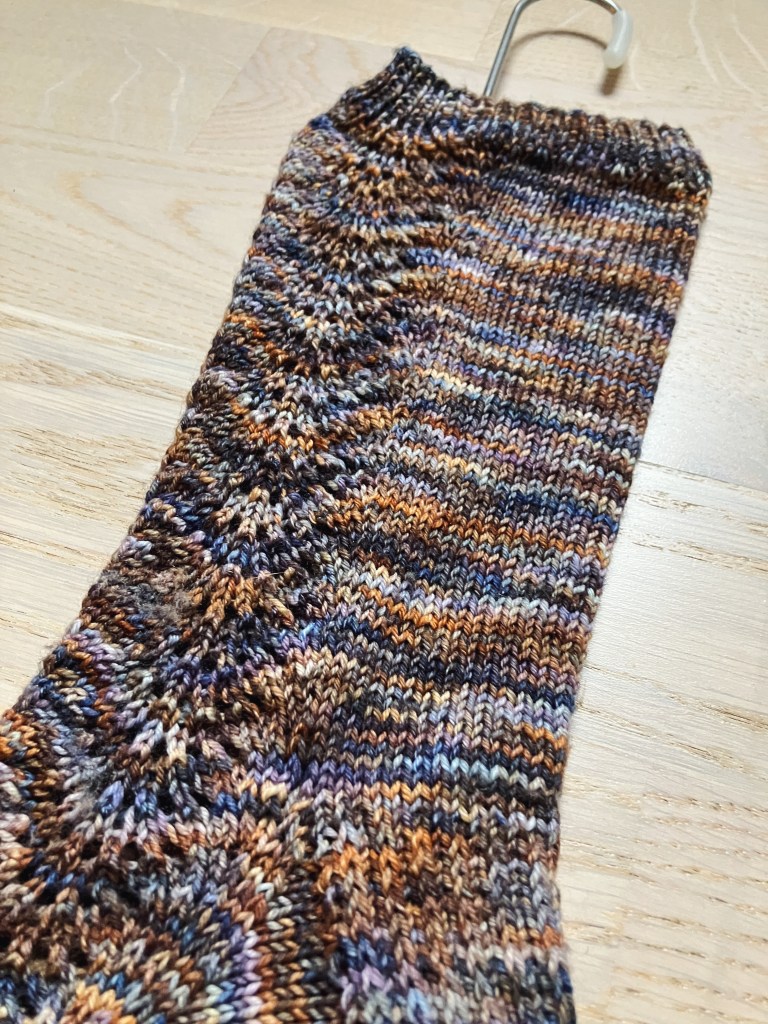 Detail of the leg of a hand-knitted sock. The left side features feather and fan lace while the right half is stockinette. The hand-dyed colorway features deepy bluey-purples, browns, and oranges in a variegated pattern.